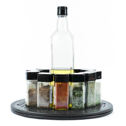 A clear glass bottle of oil stands upright in the center of a circular black tray, surrounded by eight small clear spice or herb jars with black lids, all arranged on a white background.