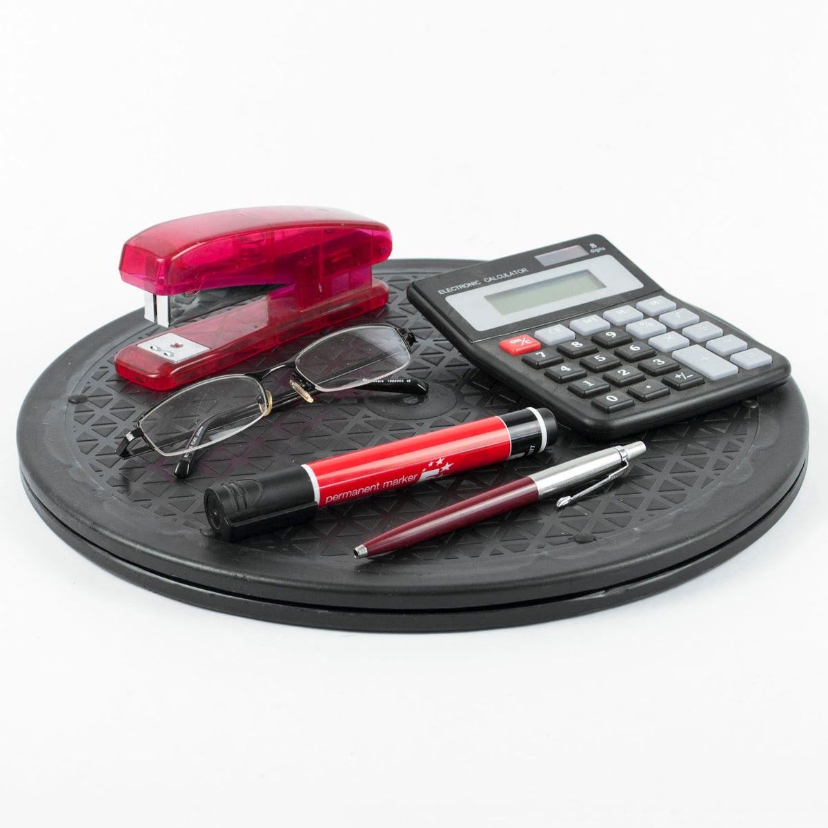 A desk accessory set arranged on a round black rotating platform against a white background. The set includes a translucent red plastic stapler, a black calculator, a pair of eyeglasses, a red and black ballpoint pen, and a metallic burgundy and silver rollerball pen.