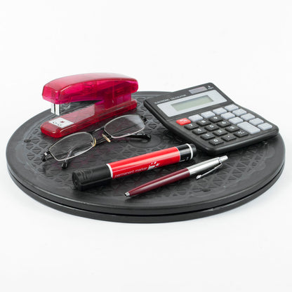 A desk accessory set arranged on a round black rotating platform against a white background. The set includes a translucent red plastic stapler, a black calculator, a pair of eyeglasses, a red and black ballpoint pen, and a metallic burgundy and silver rollerball pen.