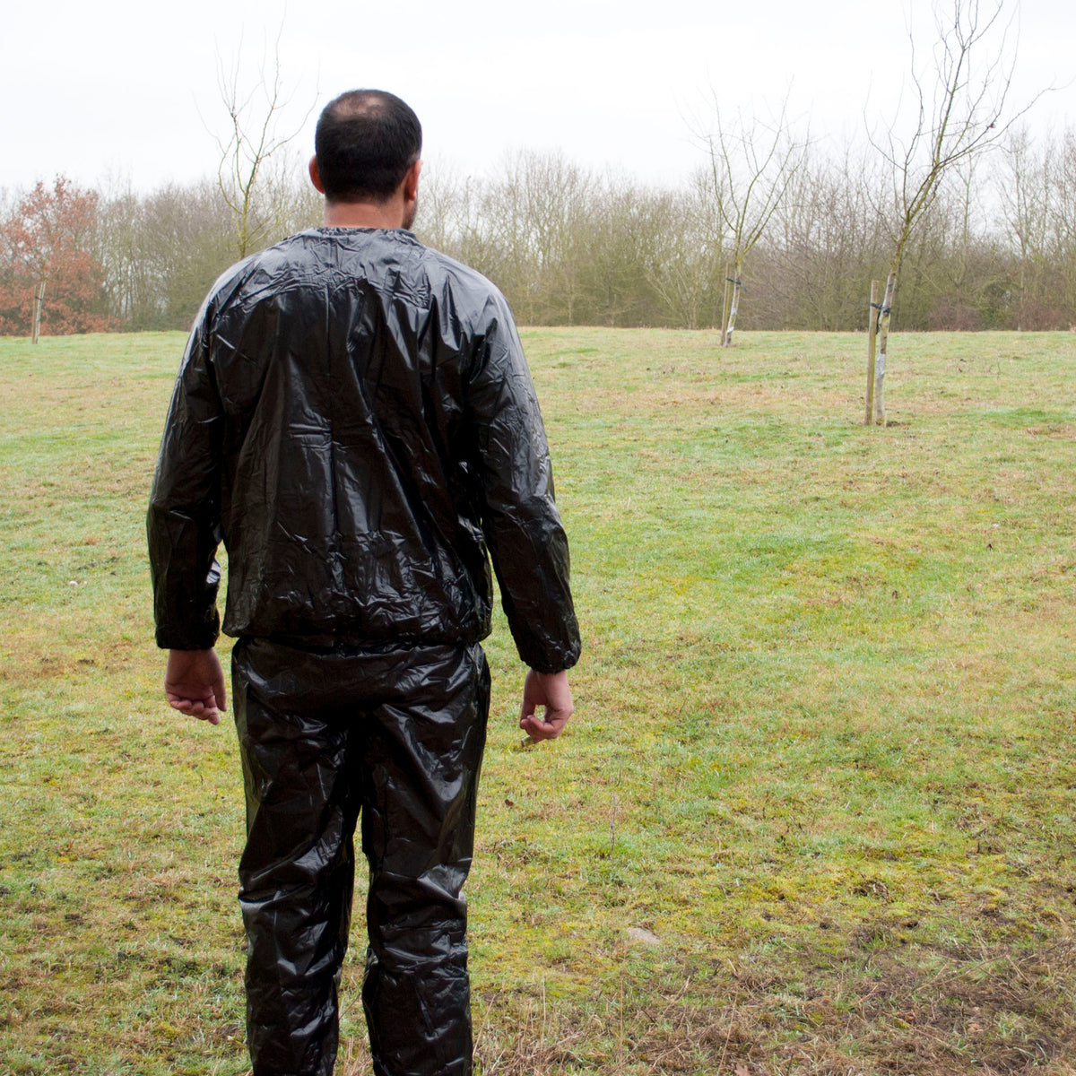 A man viewed from behind wearing a shiny black sauna suit, standing in a grassy field with bare trees in the background.