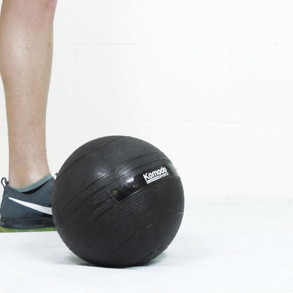 A black Komodo slam ball on a white floor next to a person's leg and shoe.