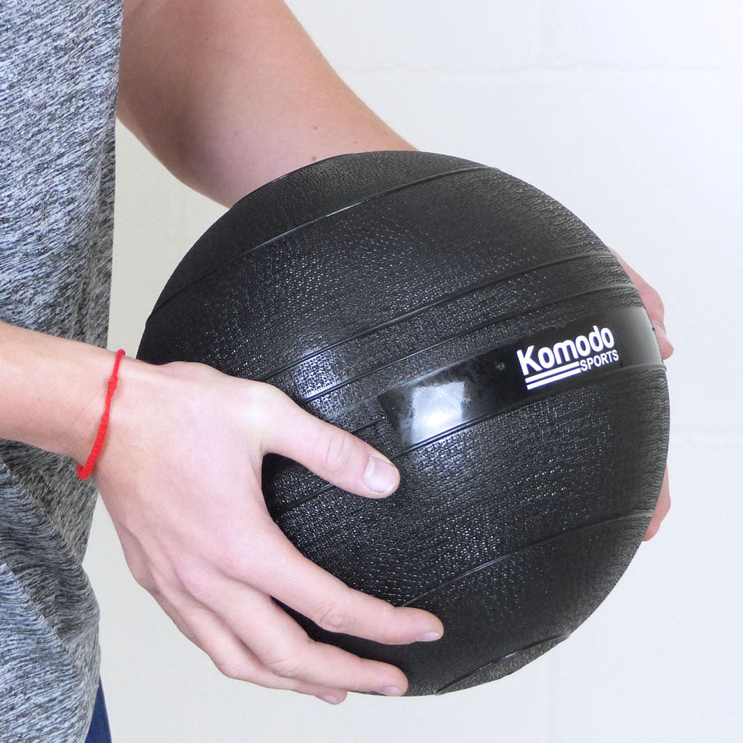 A person holding a black textured Komodo Sports slam ball against a light background.