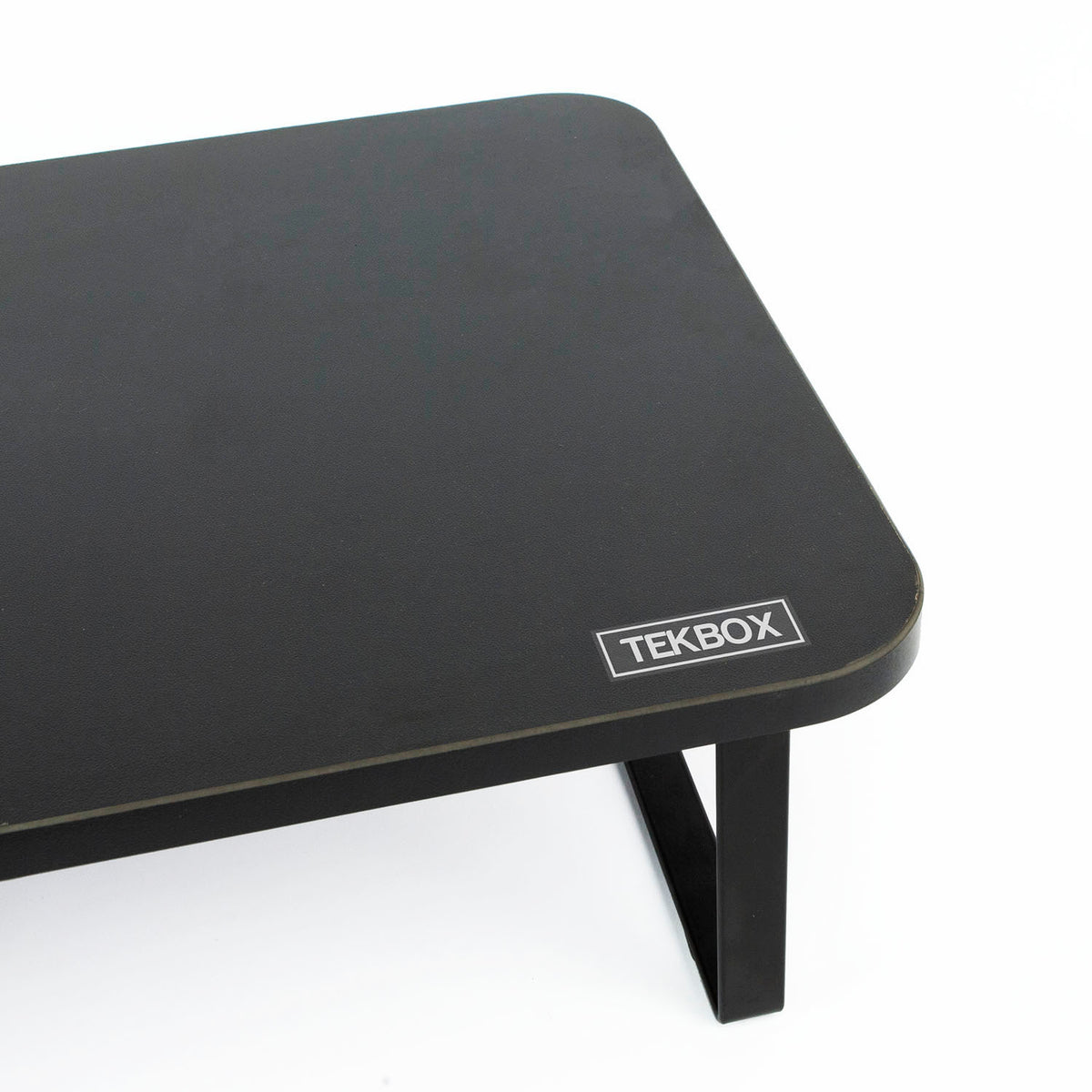 A close-up of a black rectangular tabletop with rounded corners and a visible TEKBOX logo badge on the front edge, set against a plain white background.
