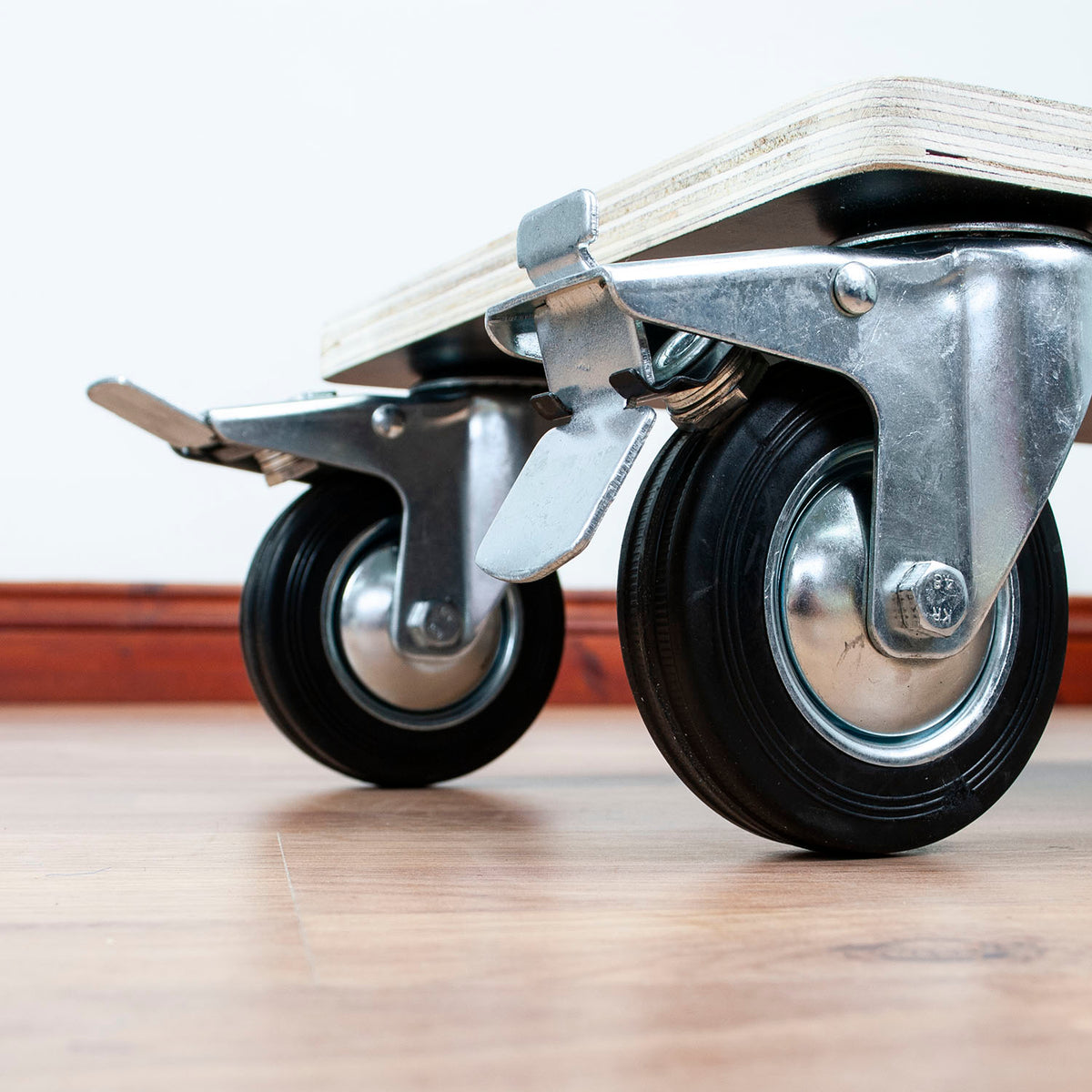 Close-up of the casters and underside of a wooden rolling board or dolly on a wood floor against a white wall.