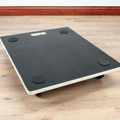 A black transport board with four circular non-slip feet and a white wood edge, shown on a light wood floor in an indoor room setting.
