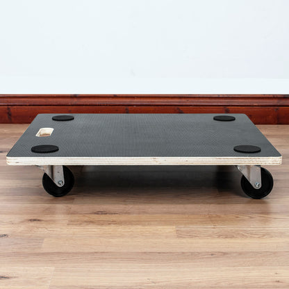 A four-wheel dolly cart with a black textured rubber mat, light wood edges, and black caster wheels, shown on a light wood floor against a white wall.