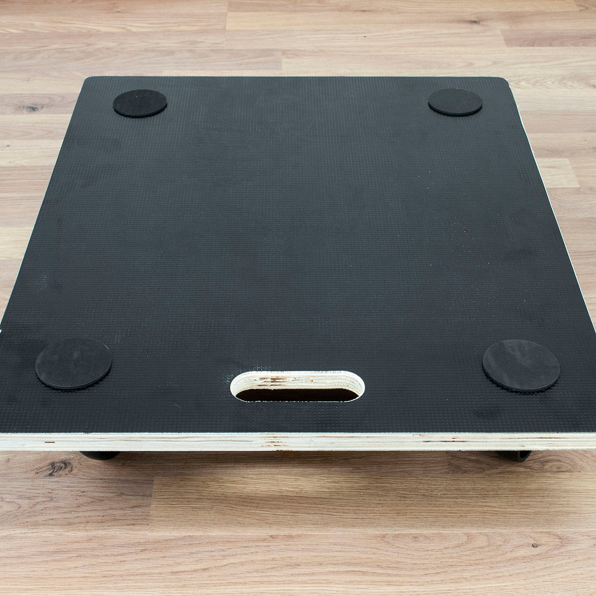 Black textured platform board with four circular rubber feet and a central cutout, shown on a light wood floor.
