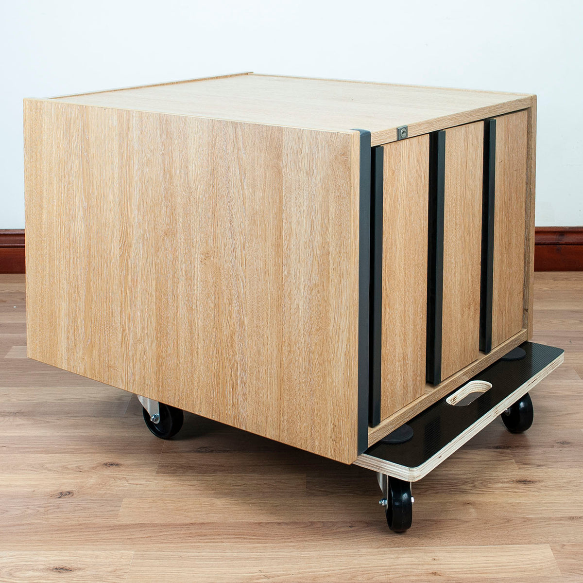 A light wood rolling storage cabinet with black metal frame and caster wheels, shown on a wood floor against a white wall.