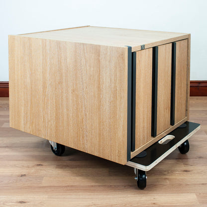 A light wood rolling storage cabinet with black metal frame and caster wheels, shown on a wood floor against a white wall.