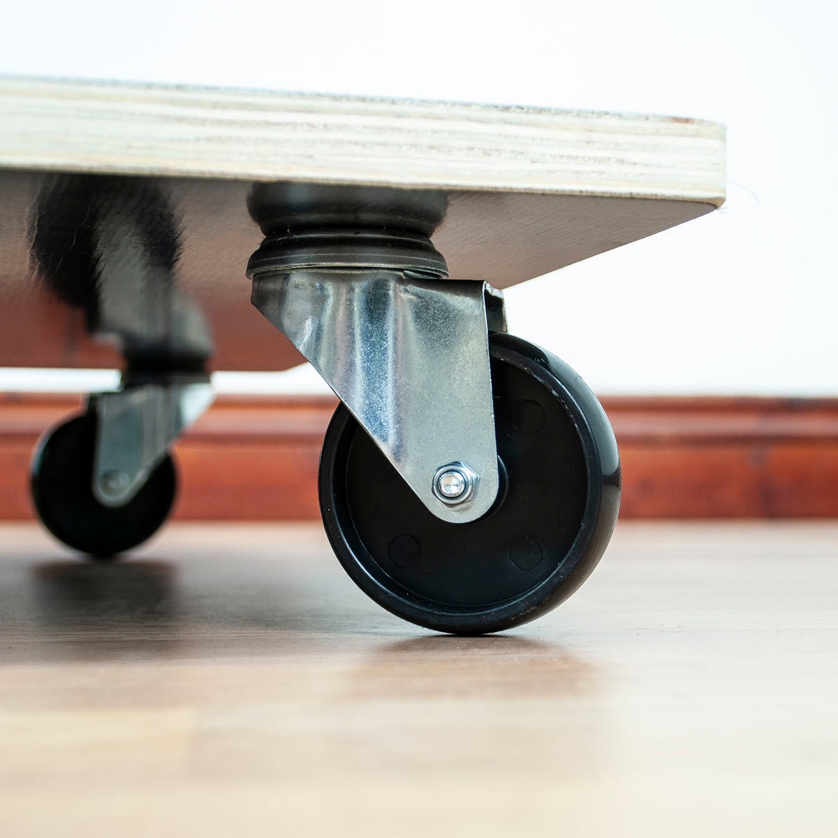Close-up of a black caster wheel with a metal swivel bracket attached to the underside of a light-colored wooden platform, set on a wooden floor.