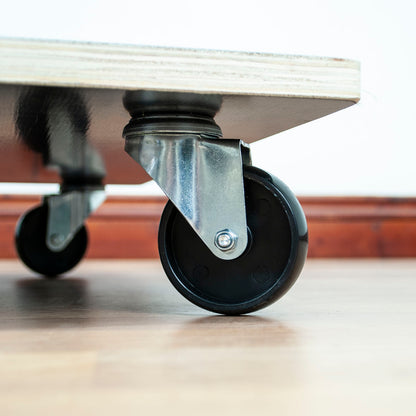 Close-up of a black caster wheel with a metal swivel bracket attached to the underside of a light-colored wooden platform, set on a wooden floor.
