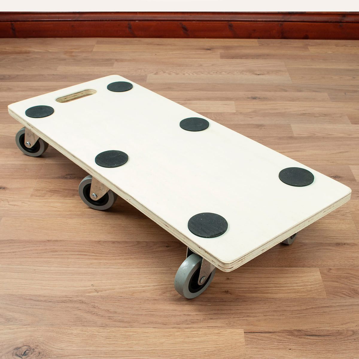 A light-colored wooden dolly with six gray caster wheels and black rubber pads, photographed on a wooden floor.