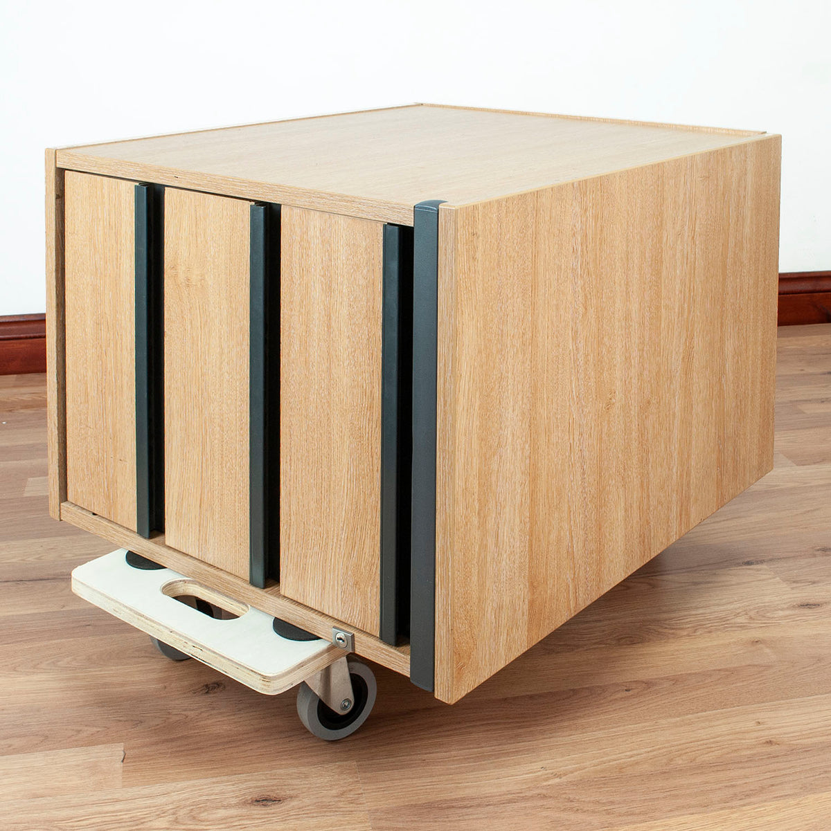 A light wood rolling storage cabinet with three front doors, dark metal handles, a wooden step platform, and caster wheels, shown on a wood floor against a white wall.