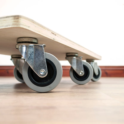 Close-up of a light wood board with four gray caster wheels on a wooden floor against a white wall.