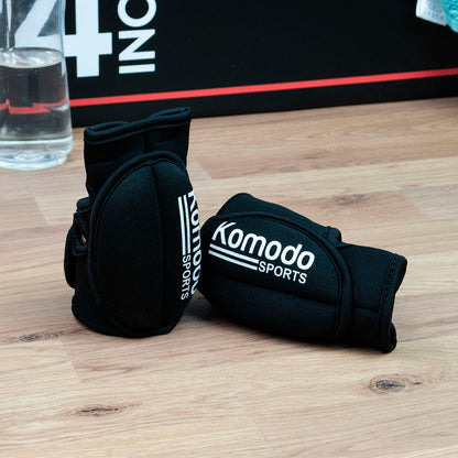 Black A black neoprene fingerless hand grip/weightlifting gloves worn on a person's hand, featuring the white 'Komodo Sports' logo printed on the back, set against a plain white background. on a wooden surface.