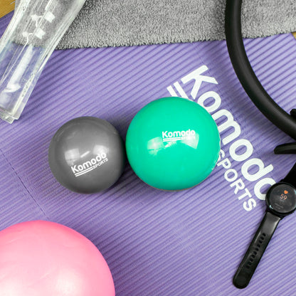 Two Komodo Sports exercise balls, one gray and one mint green, placed on a purple Komodo Sports mat alongside fitness accessories including a pink ball, smartwatch, and a clear water bottle.
