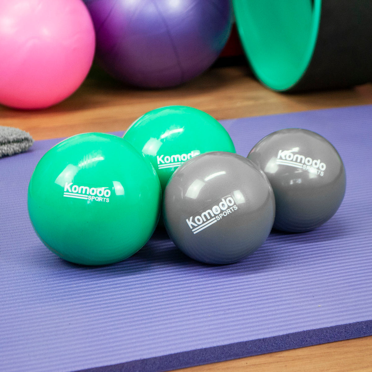Four Komodo Sports Weighted Toning Ball in green and grey arranged on a purple yoga mat, with colorful exercise balls in the background on a wooden surface.