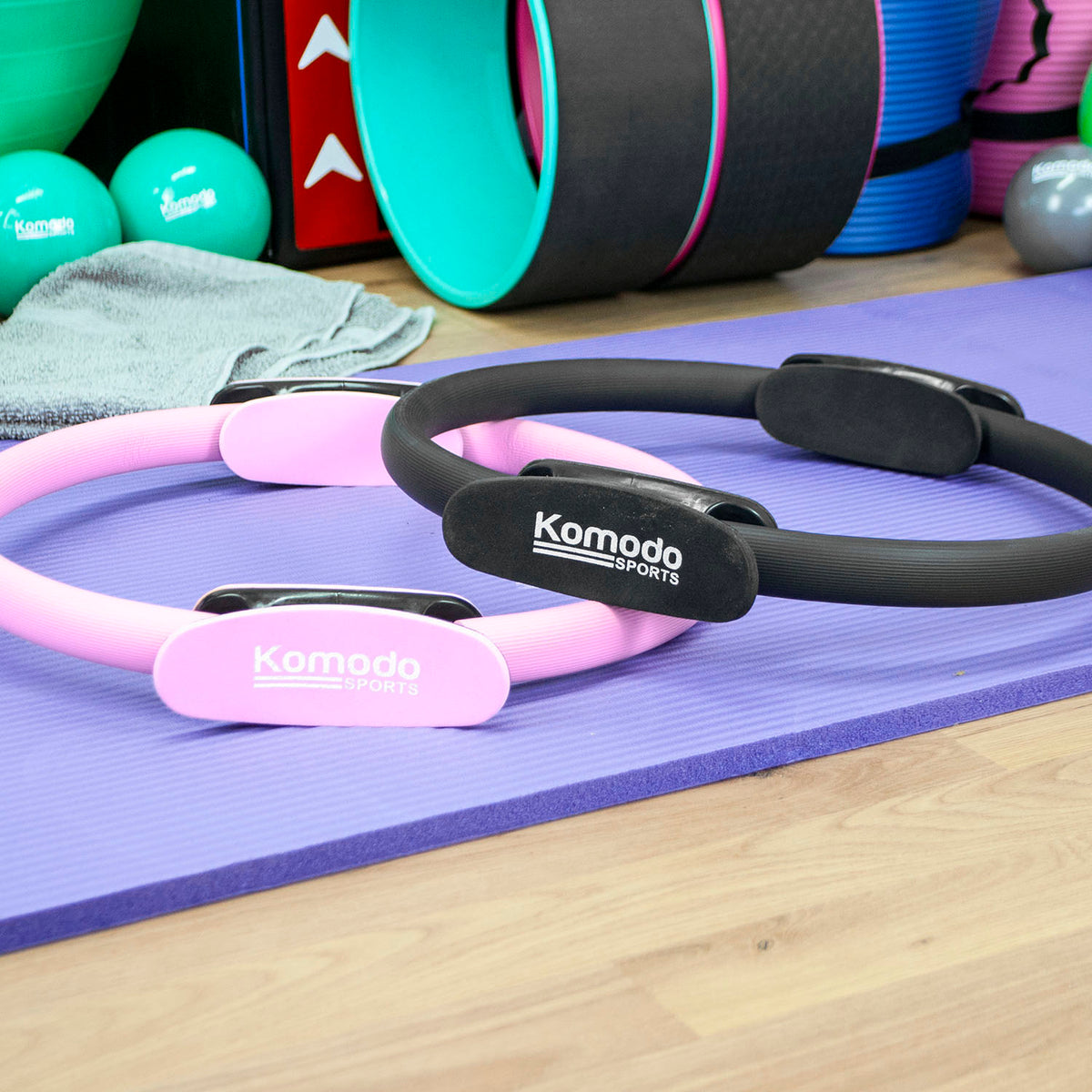 Two Komodo Sports Pilates reformer rings, one pink and one black, resting on a purple yoga mat in a gym setting with exercise equipment in the background.