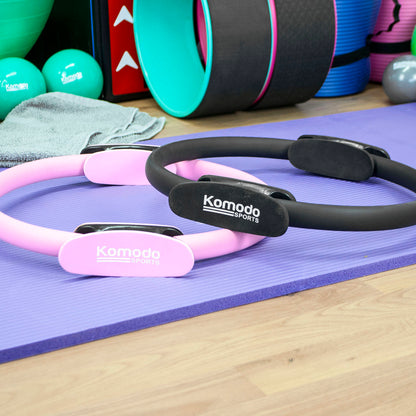 Two Komodo Sports Pilates reformer rings, one pink and one black, resting on a purple yoga mat in a gym setting with exercise equipment in the background.